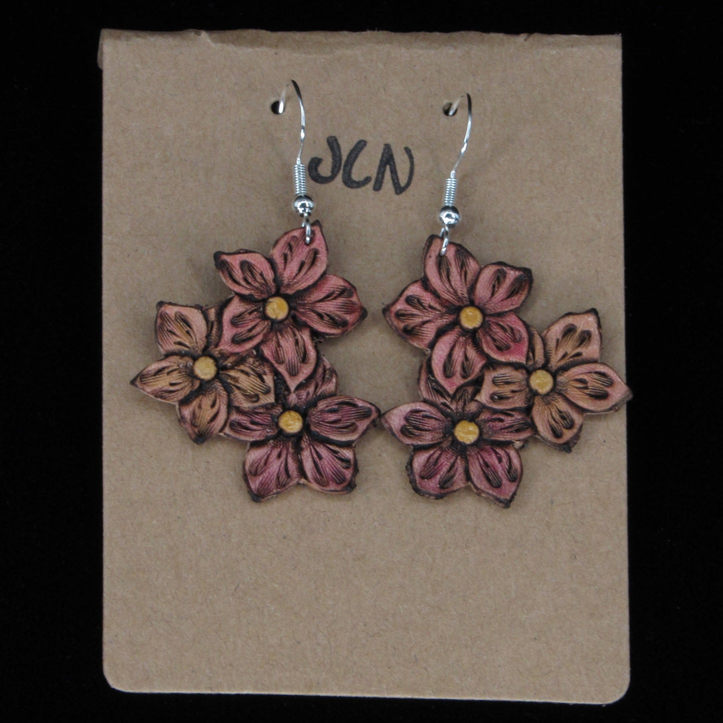 Flower-shaped leather earrings on a brown card with 'JLN' branding against a black background