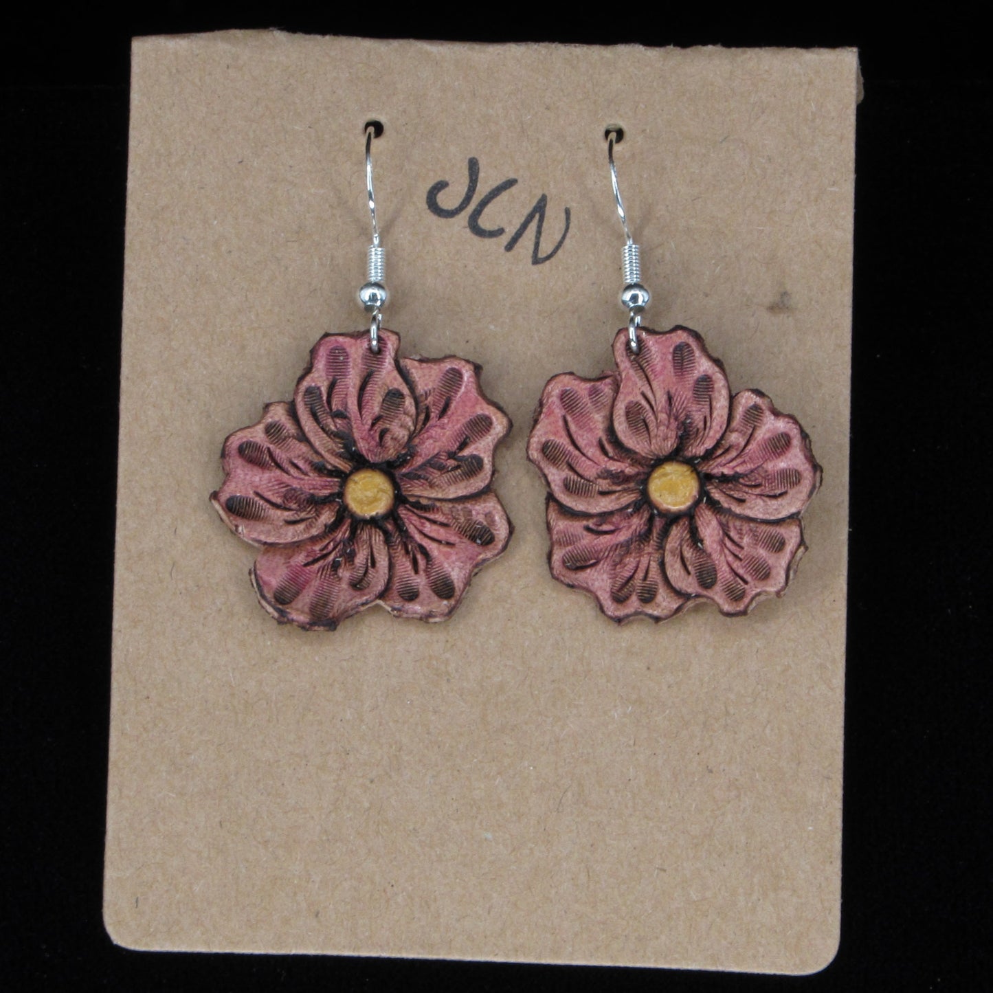 Flower-shaped leather earrings on a brown card with 'JLN' branding against a black background