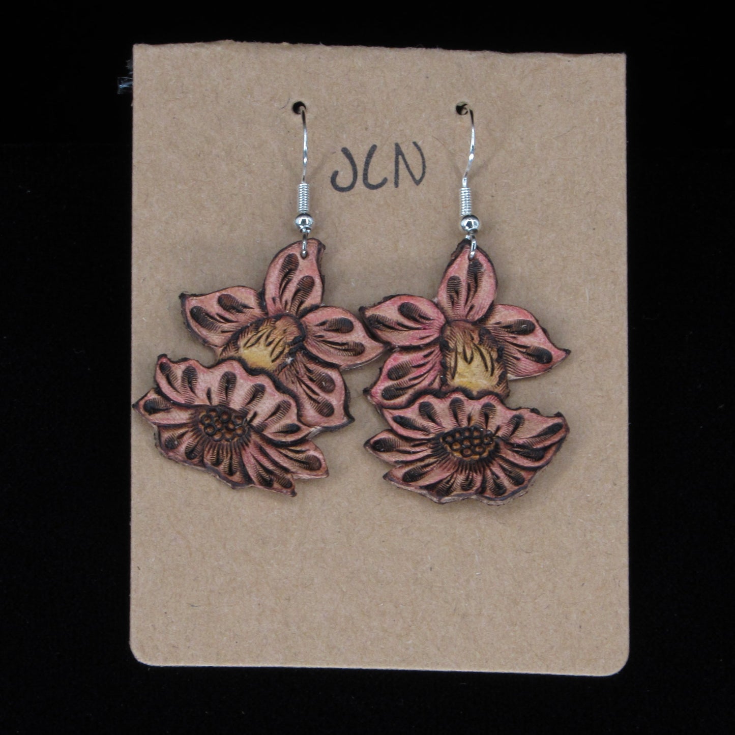 Flower-shaped leather earrings on a brown card with 'JLN' branding against a black background