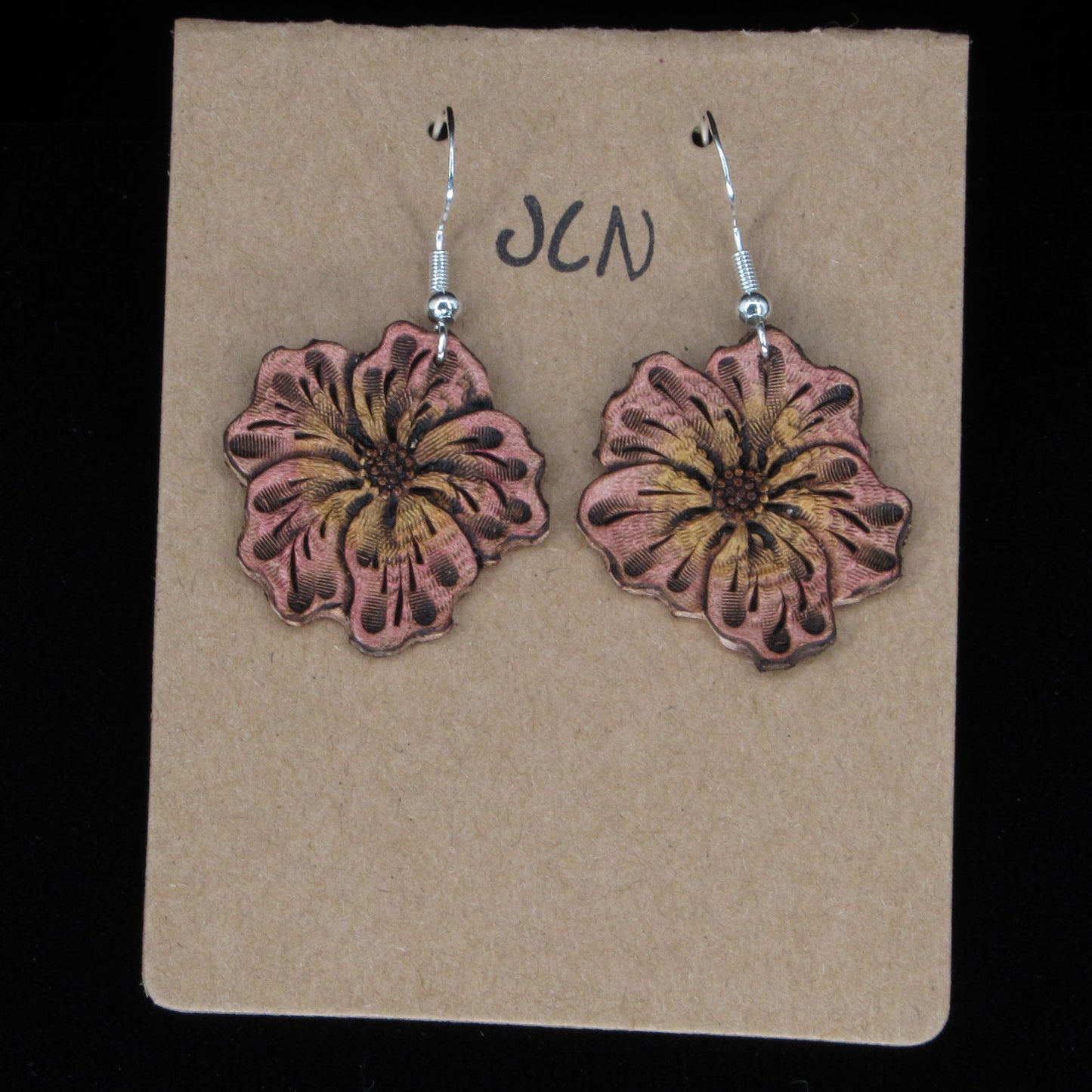 Pink flower-shaped earrings on a beige card with 'JLN' branding against a black background