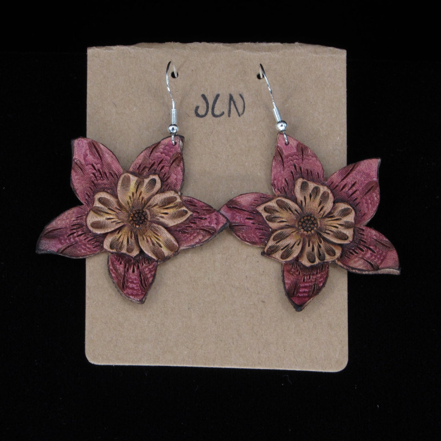 Flower-shaped leather earrings on a brown card with 'JLN' branding against a black background