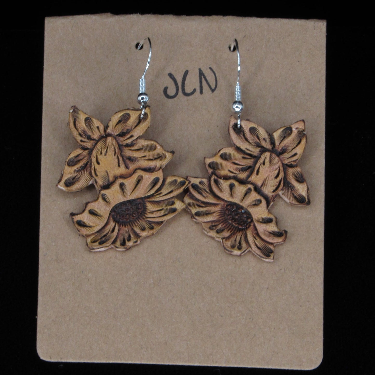 Flower-shaped leather earrings on a brown card with 'JLN' branding against a black background