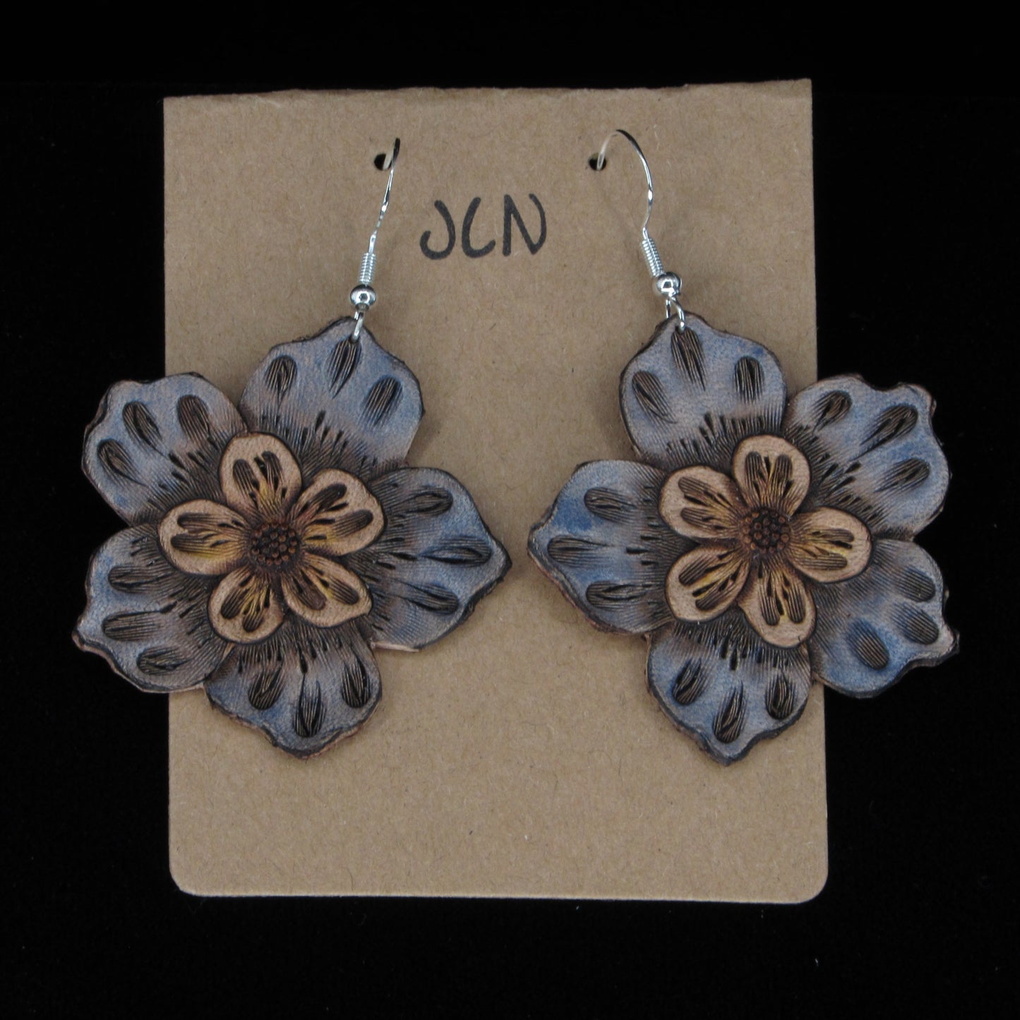 Blue flower-shaped earrings on a beige card with 'JLN' branding against a black background