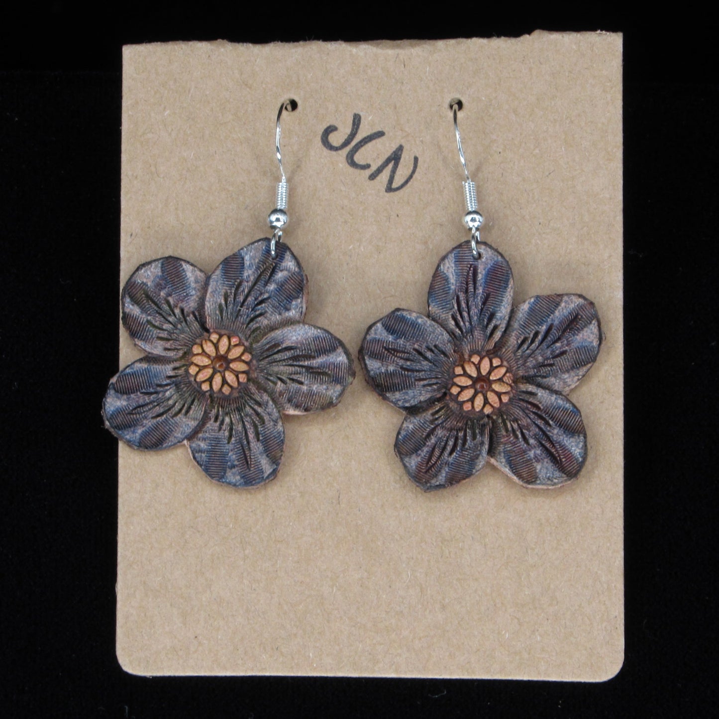 Flower-shaped leather earrings on a brown card with 'JLN' branding against a black background